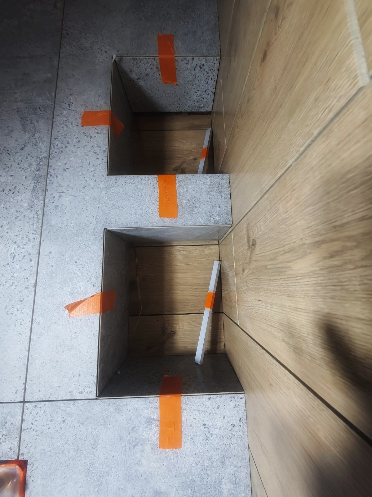 Przygotowanie do ułożenia płytek w narożniku z drewnopodobną okładziną ścienną, widoczne pomarańczowe taśmy maskujące i listwy dystansowe.