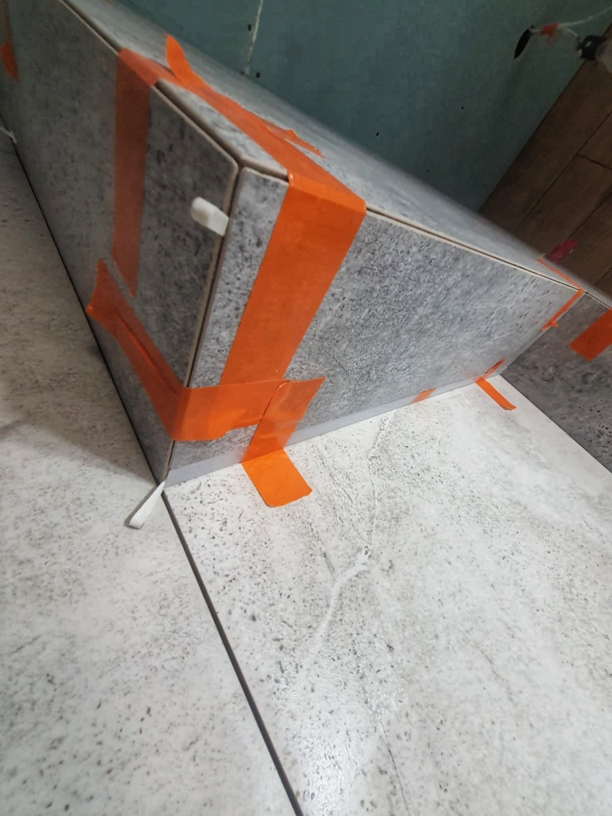 Szara płytka imitująca kamień, ułożona na schodku, zabezpieczona pomarańczową taśmą malarską i plastikowymi krzyżykami dystansowymi.
