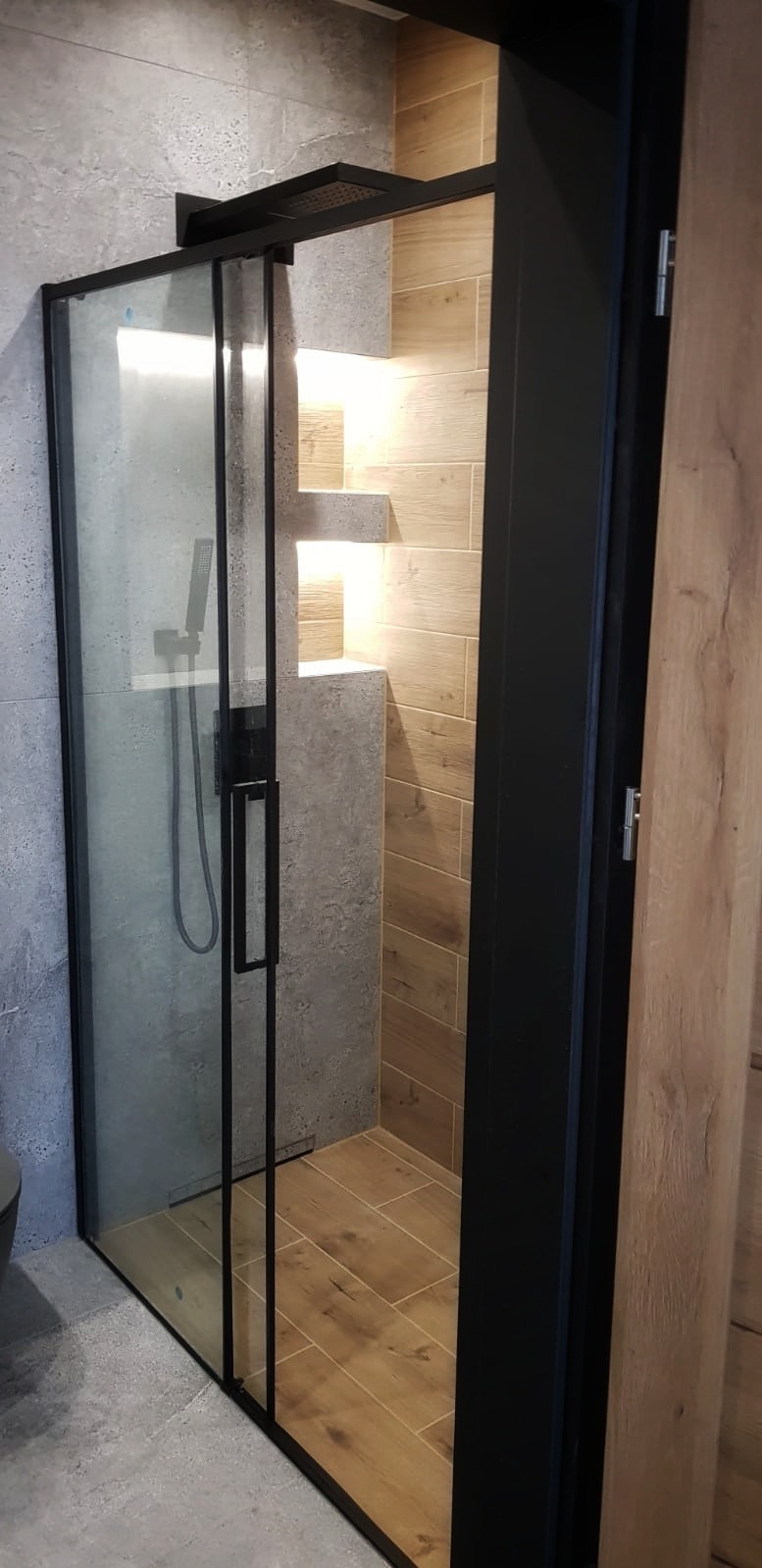 Nowoczesna kabina prysznicowa z czarnymi profilami i deszczownicą w wykończonej łazience w Gliwicach. Widoczne szare płytki na ścianie i podłodze oraz drewnopodobne płytki wewnątrz kabiny...