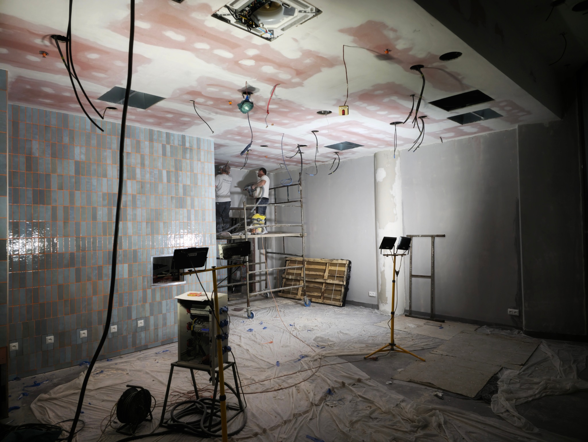 Prace wykończeniowe w pomieszczeniu: robotnicy na rusztowaniu montują instalację elektryczną pod sufitem z widocznymi śladami szpachlowania, ściana wyłożona pionowymi, niebieskimi płytkami...