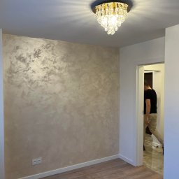 DaMa Bud - Ściana w pokoju wykończona dekoracyjnym tynkiem w odcieniach beżu i srebra, z widocznym kryształowym żyrandolem i fragmentem osoby przechodzącej przez drzwi.
