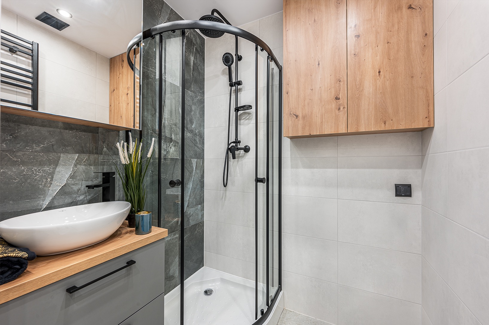 Nowoczesna łazienka z kabiną prysznicową z czarnymi profilami, umywalką nablatową na drewnianym blacie i szarą szafką, drewniane szafki wiszące, szare i białe płytki na ścianach.