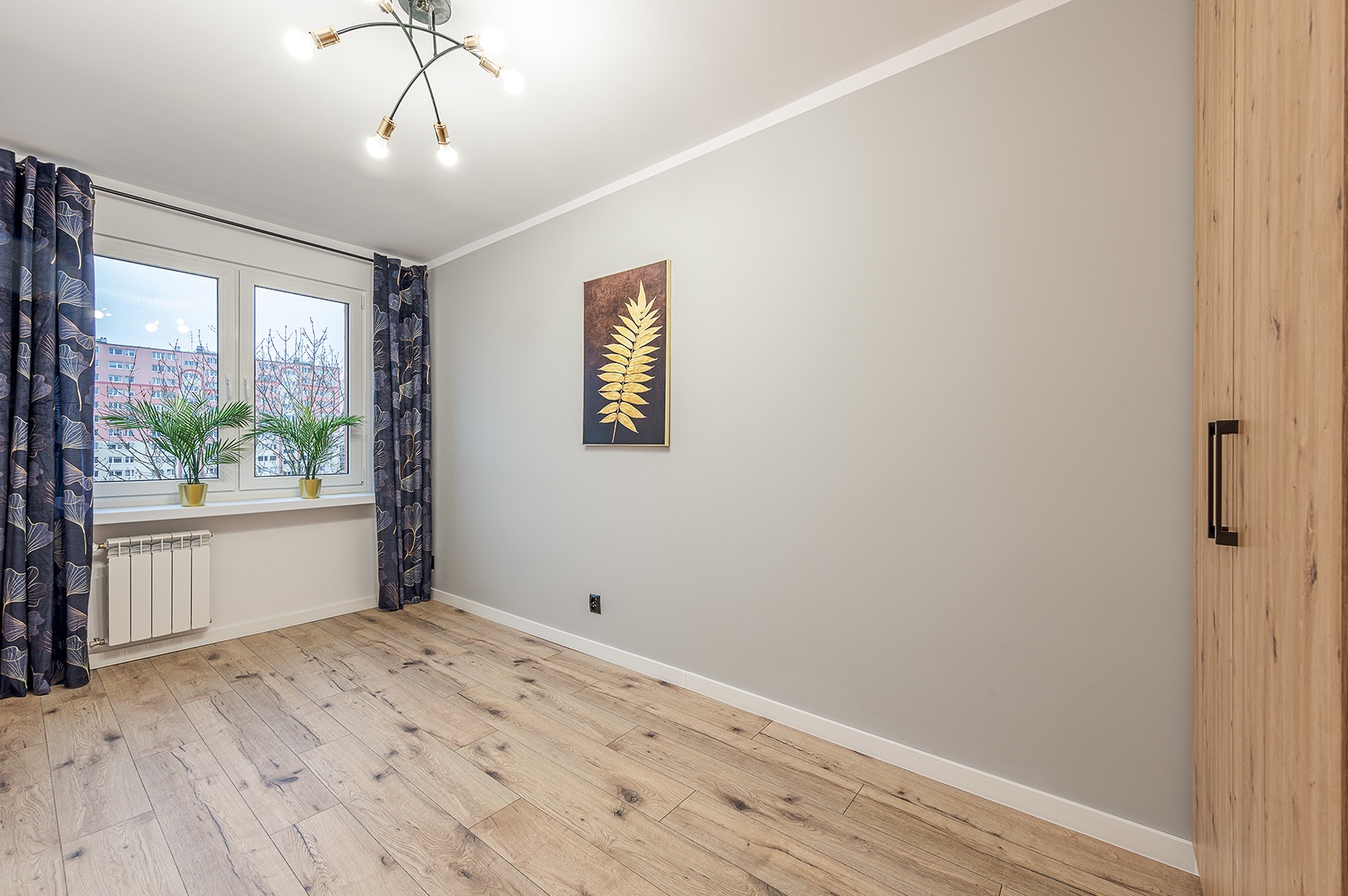 Pusty pokój po odświeżeniu: jasne panele podłogowe, szare ściany z obrazem liścia, okno z ciemnymi zasłonami i roślinami w złotych doniczkach na parapecie, biały grzejnik, drewniana szafa...