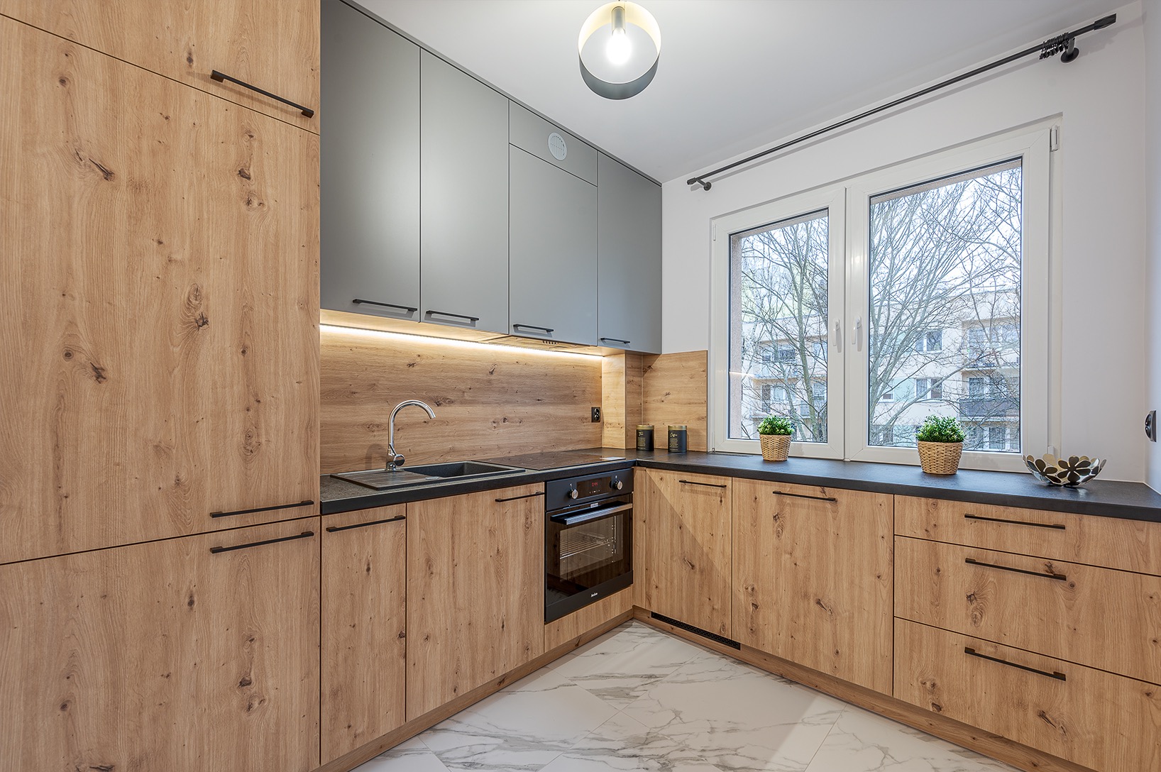 Nowoczesna kuchnia w Łodzi z drewnianymi frontami dolnych szafek i szarymi górnymi, czarnym blatem i marmurową podłogą, oświetlona lampą sufitową i światłem spod szafek.