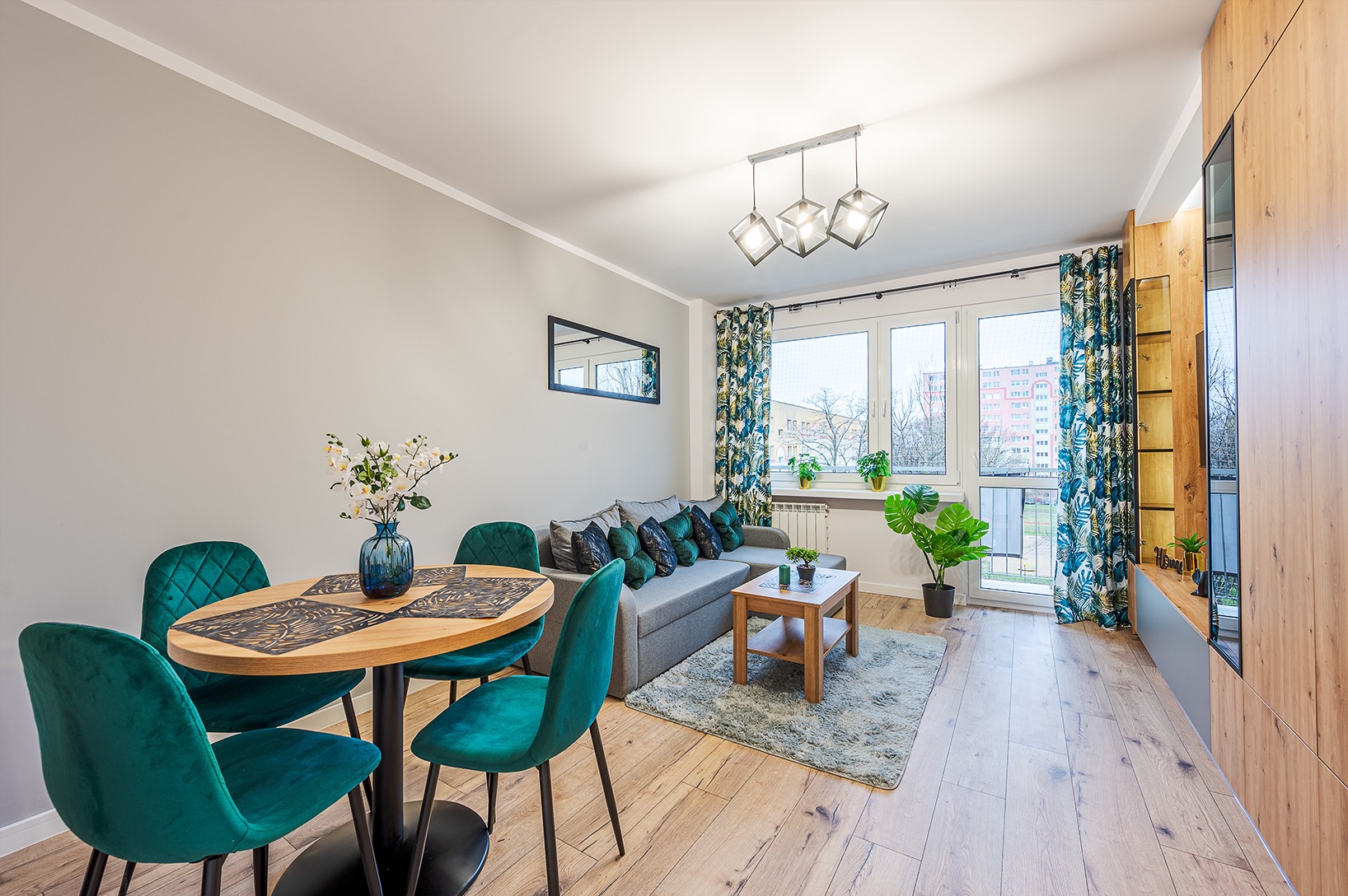 Jasne wnętrze mieszkania z drewnianą podłogą, okrągłym stołem z zielonymi krzesłami, szarą sofą i drewnianą zabudową ściany, doświetlone nowoczesną lampą wiszącą.