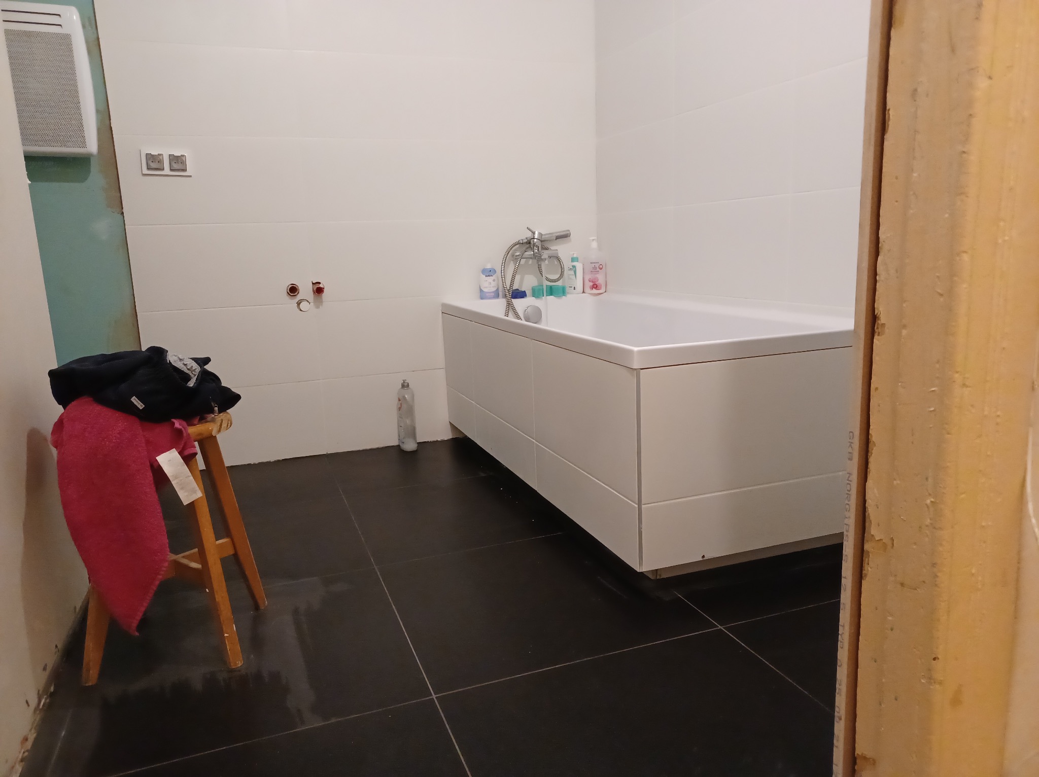 Biała wanna obudowana płytami w trakcie montażu, widoczne niezakończone prace wykończeniowe, czarne płytki podłogowe, drewniany taboret z ubraniami i ręcznikiem oraz wystające elementy instalacji...