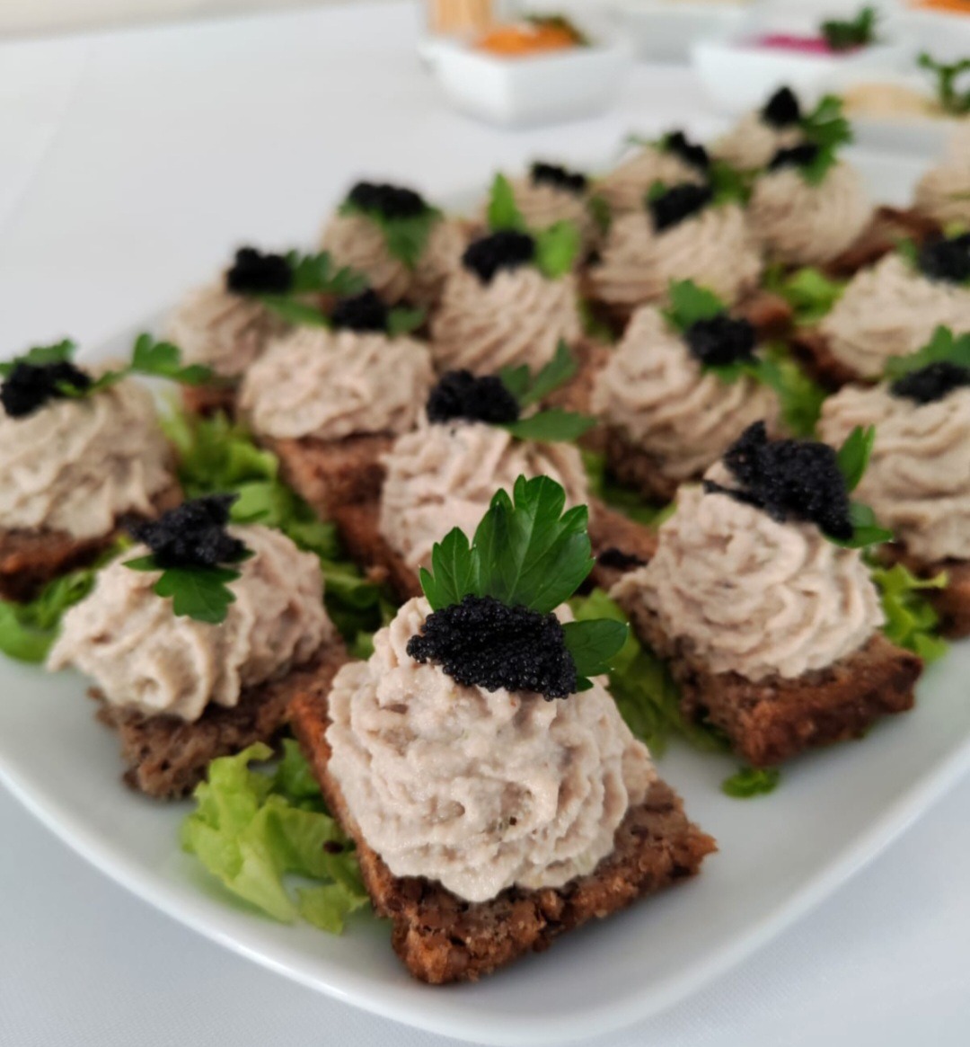 Eleganckie kanapeczki na białym talerzu, z brązowym pieczywem, kremowym musem, czarnym kawiorem i zielonymi listkami pietruszki. Idealne na przyjęcie.