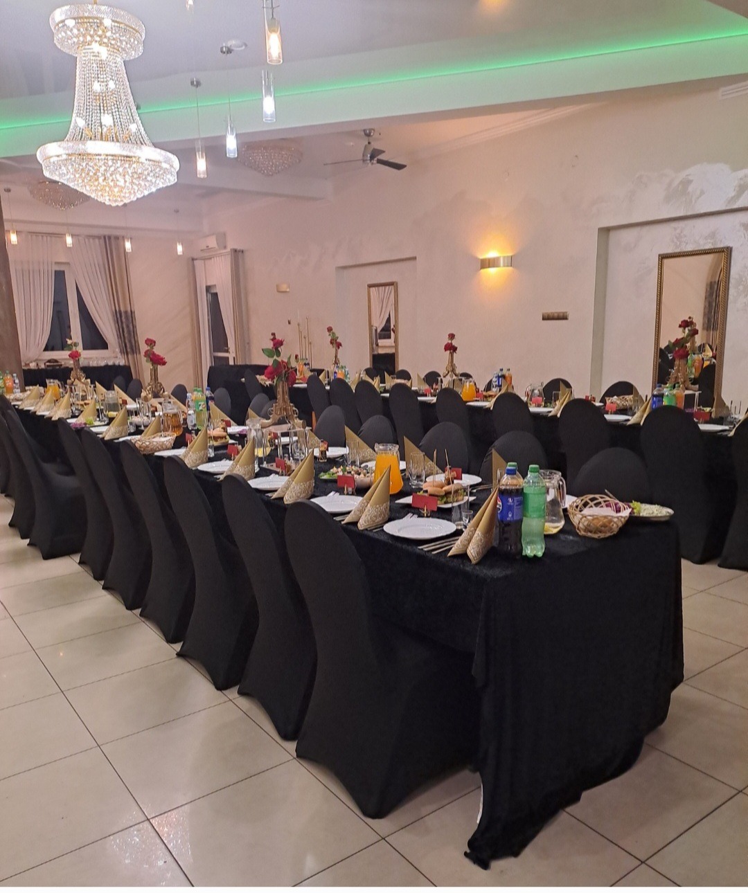 Uroczysta sala bankietowa z długim stołem, nakrytym czarnym obrusem, elegancko udekorowanym zastawą, kwiatami i napojami. Kryształowy żyrandol i zielone oświetlenie.