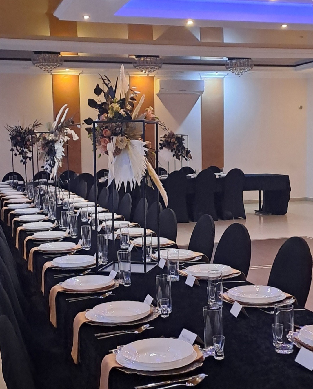 Elegancko nakryty stół na przyjęcie z czarnym obrusem, białymi talerzami, złotymi sztućcami i wysokimi kompozycjami kwiatowymi w nowoczesnych, metalowych konstrukcjach.