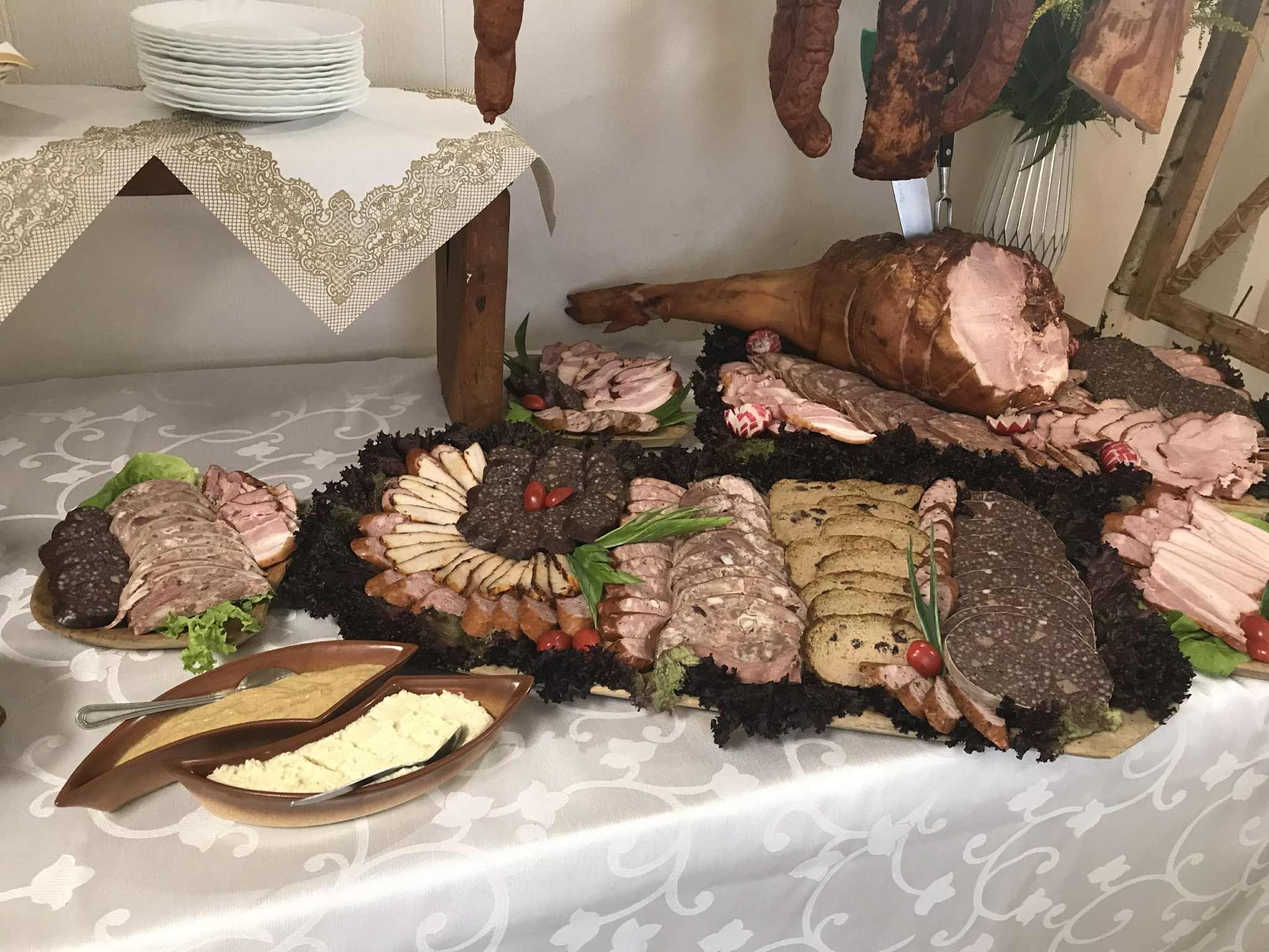 Bogato zastawiony stół z wędlinami, pasztetami i sosami na białym obrusie z koronkowym wykończeniem. W tle wiszące wędzonki i stos talerzy.