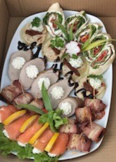 Elegancka kompozycja przekąsek na białym talerzu: roladki z tortilli, kanapeczki, wędlina z kremem, łosoś z cytryną i bekon. Idealne na firmowe przyjęcie.