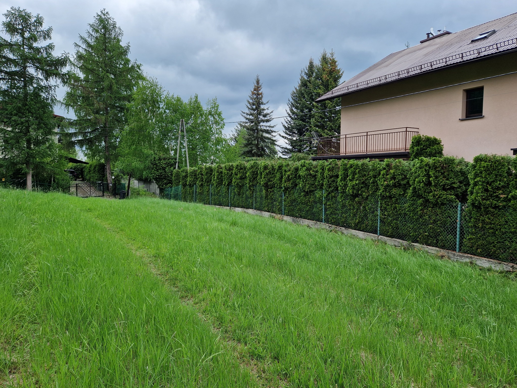 Zadbany ogród z równo przyciętym żywopłotem i wysoką trawą. W tle dom z beżową elewacją i brązowym balkonem. Pochmurne niebo nadaje spokojny charakter.