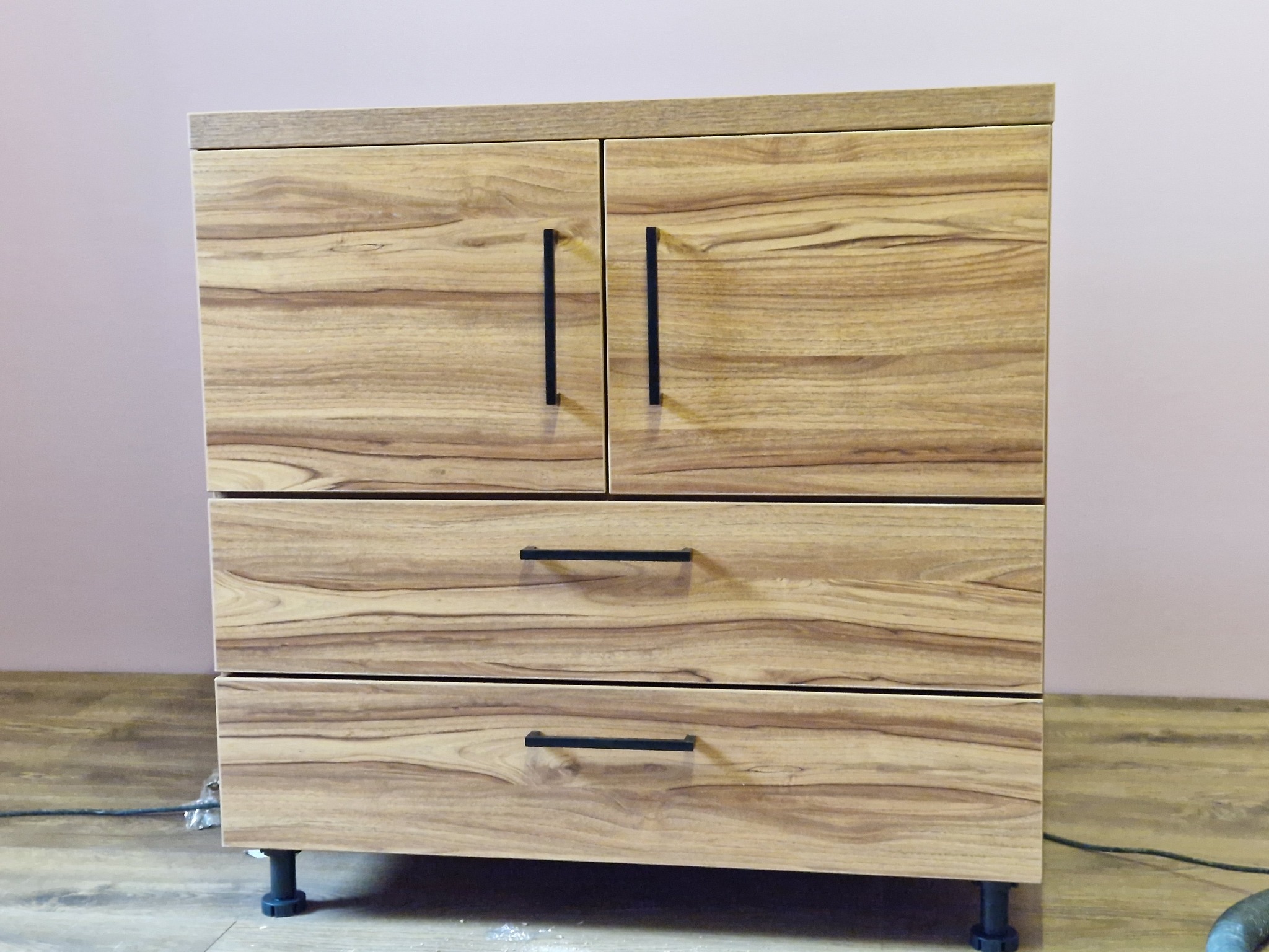 Komoda z drewna o naturalnym wzorze słojów, z dwoma szufladami i drzwiczkami. Czarne, minimalistyczne uchwyty. Ustawiona na regulowanych nóżkach.