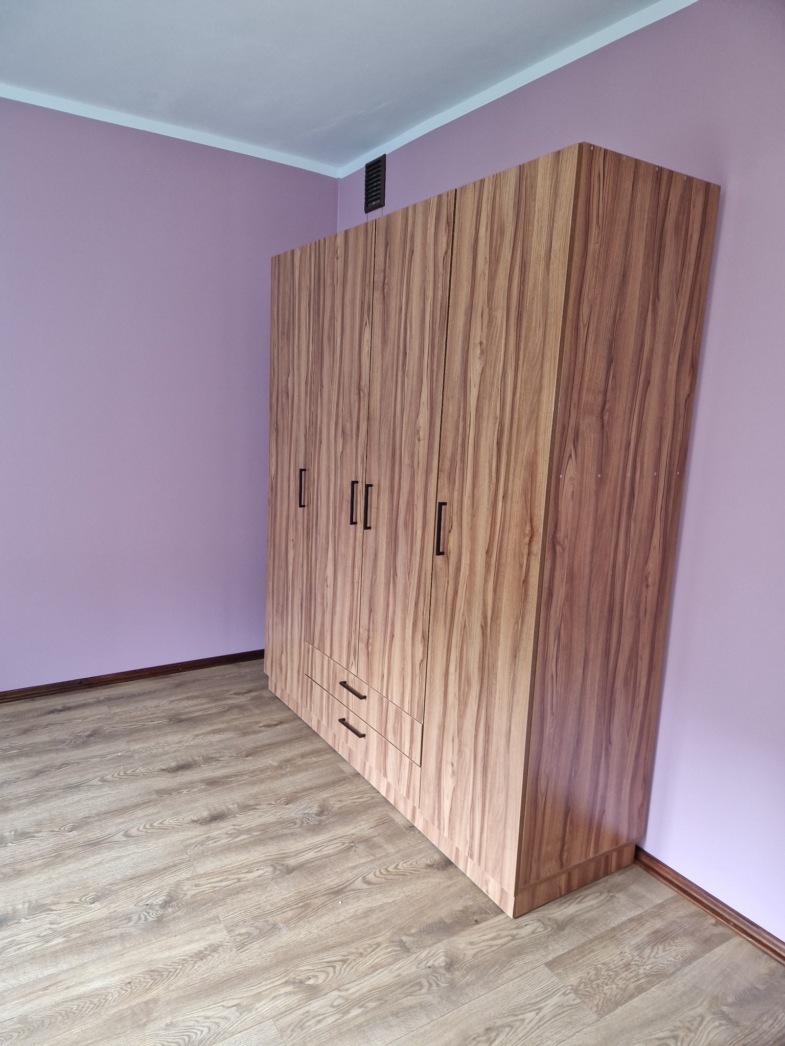 Wysoki, drewnopodobny, czterodrzwiowy szafa z dwiema szufladami, z czarnymi uchwytami, stoi na podłodze imitującej drewno, na tle liliowej ściany.