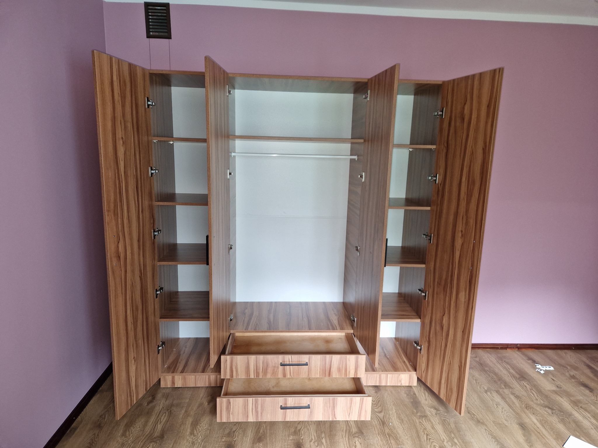 Nowoczesna, otwarta szafa z forniru w odcieniach brązu, z widocznymi półkami i szufladami, na tle fioletowej ściany. Minimalistyczny design.