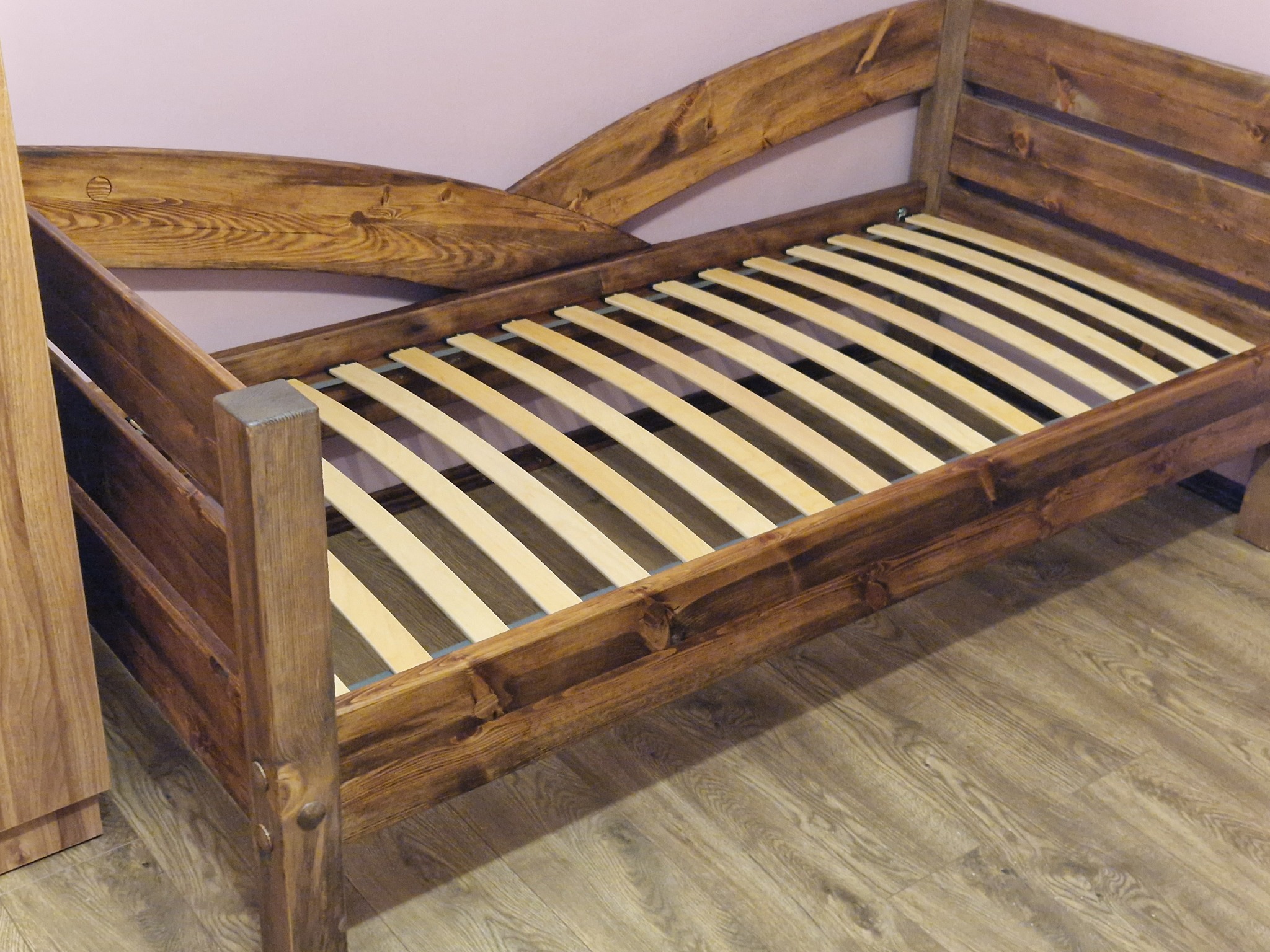 Drewniane łóżko z bejcowanymi elementami, widoczne stelaż i zagłówek o falistym kształcie. Ujęcie z perspektywy podłogi, ukazujące strukturę drewna i detale wykończenia.