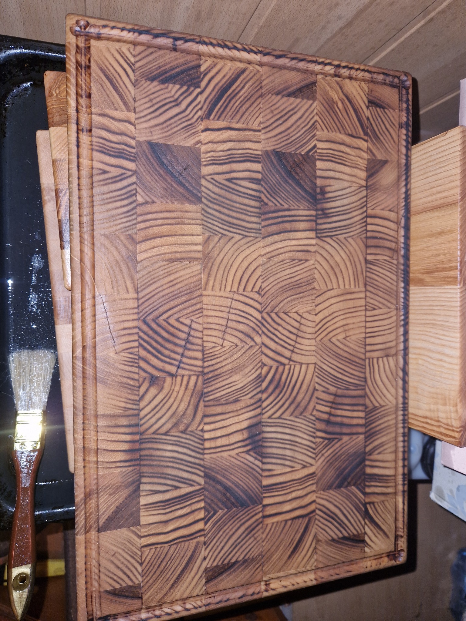 Deska do krojenia z drewna klejonego kostką, obok pędzel, na czarnej blasze. Widoczne usłojenie drewna i detale wykończenia. Stolarz.