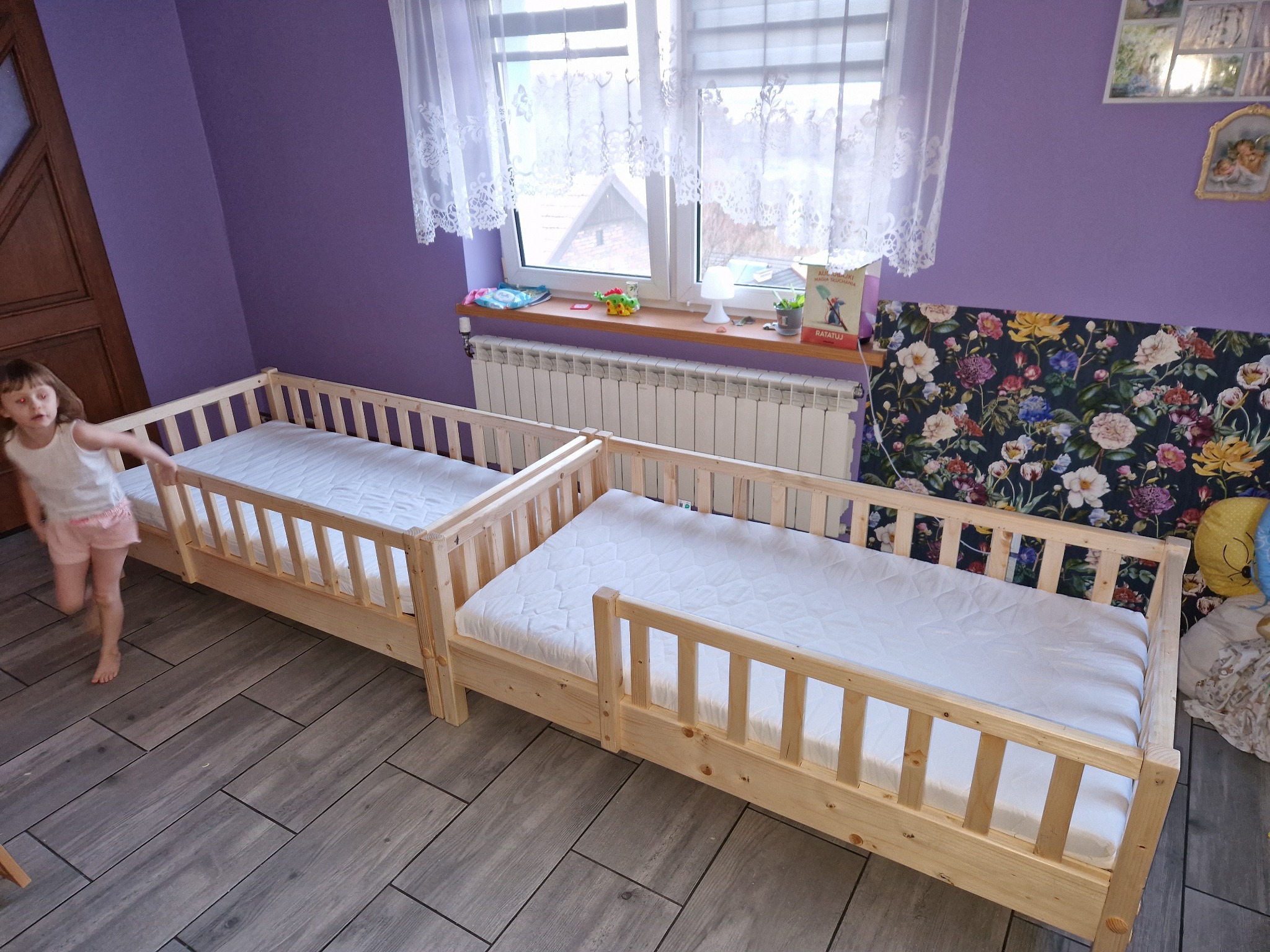 Dwa połączone drewniane łóżka dziecięce z barierkami, z białymi materacami, stoją na szarych panelach podłogowych w pokoju z fioletowymi ścianami. Dziewczynka w tle.