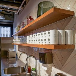 WORKSPACE-SERVICE - Biuro: Umywalka ze stali nierdzewnej, bateria, dozownik mydła i ręczników, na półkach drewnianych ułożone filiżanki i szklanki, tło stanowi ściana wyłożona błyszczącymi, kremowymi płytkami...