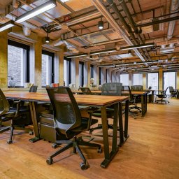 WORKSPACE-SERVICE - Przestronne biuro z drewnianą podłogą, rzędami biurek z czarnymi krzesłami, dużymi oknami i widocznymi instalacjami na suficie.