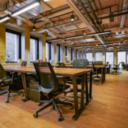 Przestronne biuro z drewnianą podłogą, rzędami biurek z czarnymi krzesłami, dużymi oknami i widocznymi instalacjami na suficie.