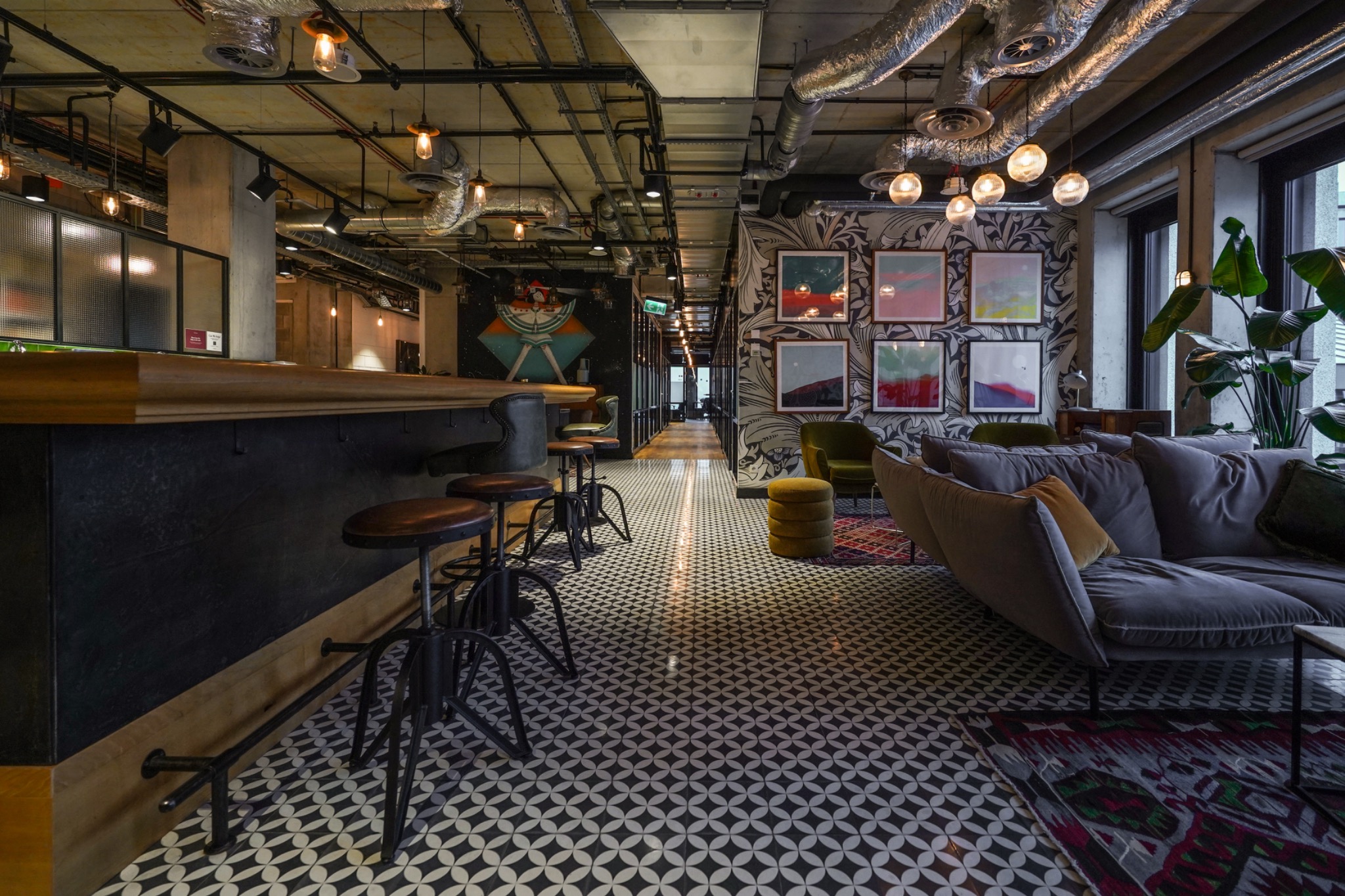 Nowoczesna przestrzeń biurowa z barem, wysokimi stołkami, wzorzystą podłogą i sofą w strefie relaksu, oświetlona wiszącymi lampami.