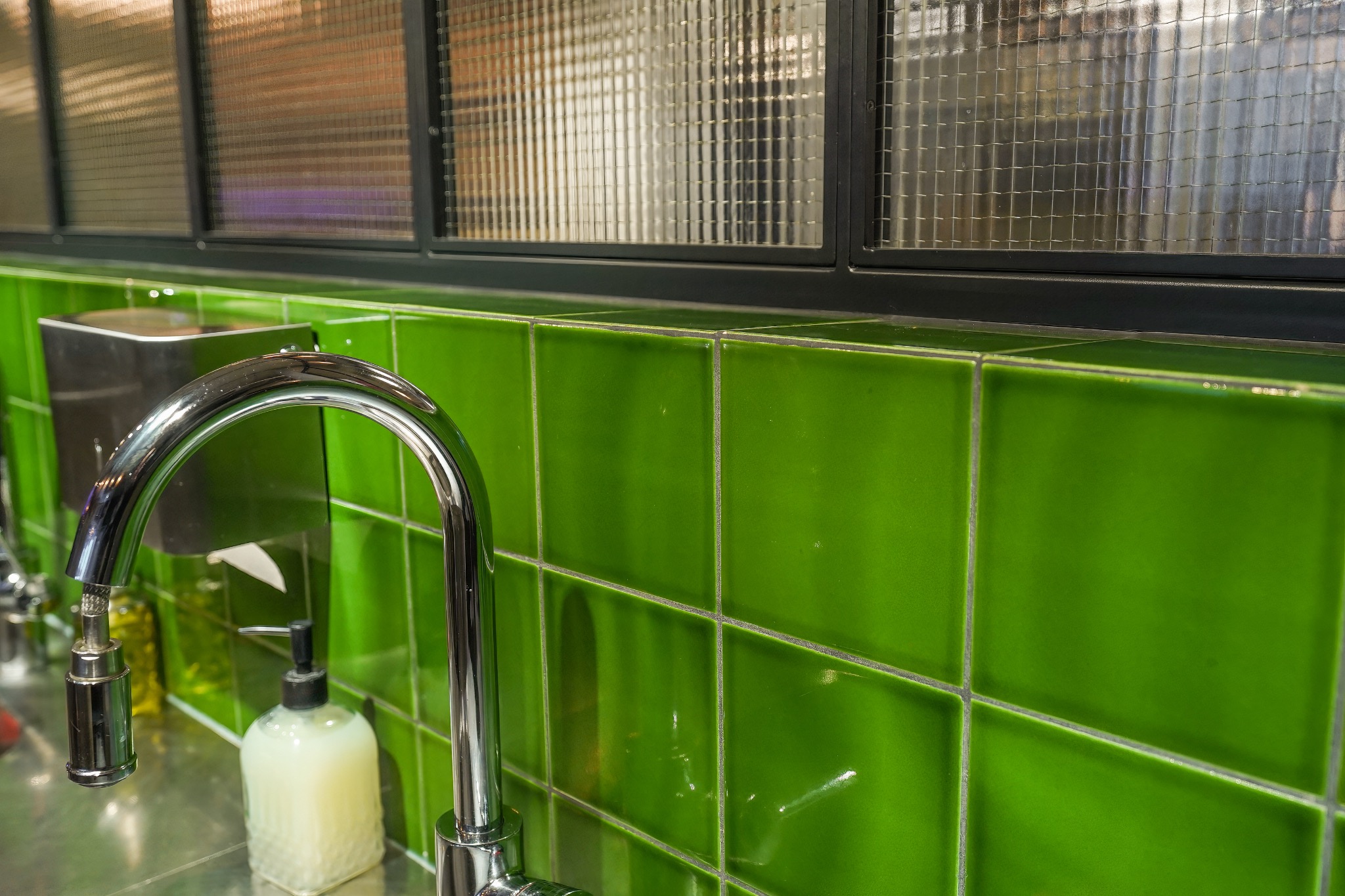 Błyszczące, zielone kafelki na ścianie w łazience w Blizne Jasińskiego, chromowana bateria umywalkowa z dozownikiem mydła w tle, okno z mlecznego szkła w czarnej ramie.
