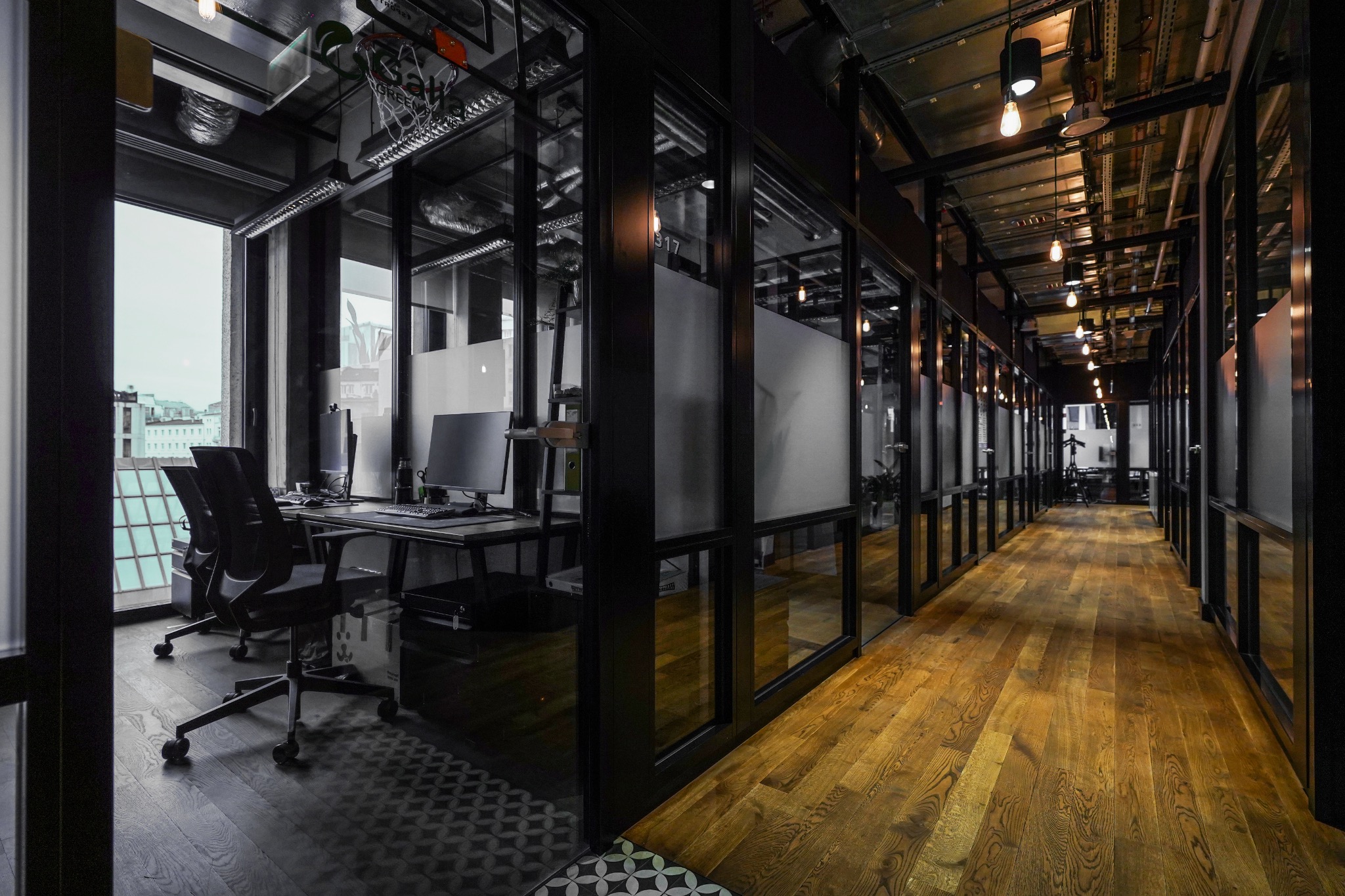Nowoczesne biuro z przeszklonymi ścianami działowymi, widok na korytarz z parkietem i stanowiska pracy z komputerami, surowy industrialny design z odsłoniętymi instalacjami pod sufitem.