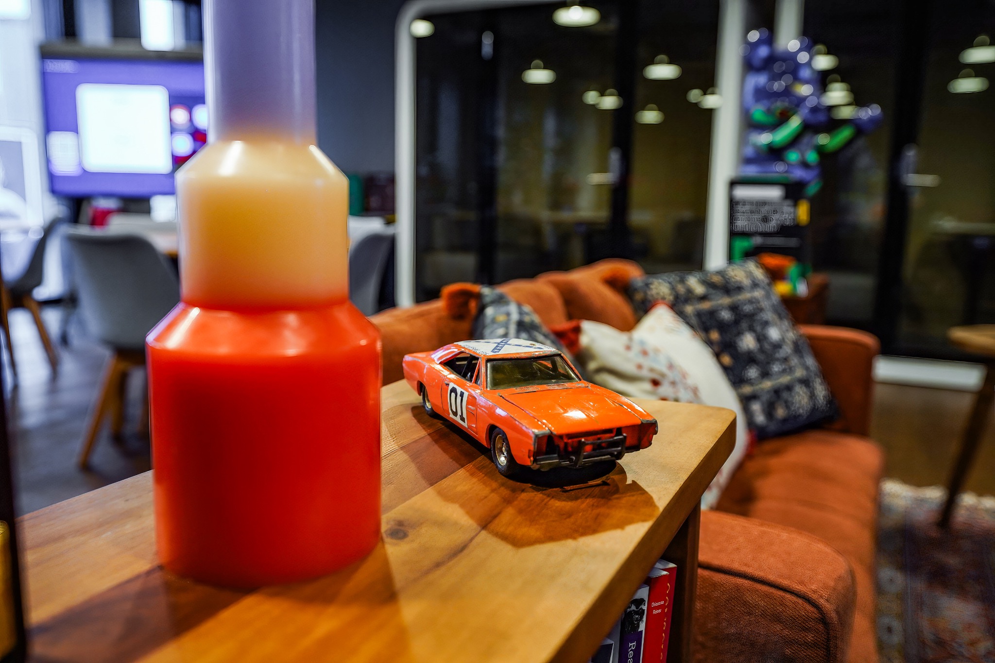 Model pomarańczowego samochodu z numerem 01 na drewnianym stoliku w biurze, obok butelki płynu, z sofą i telewizorem w tle.