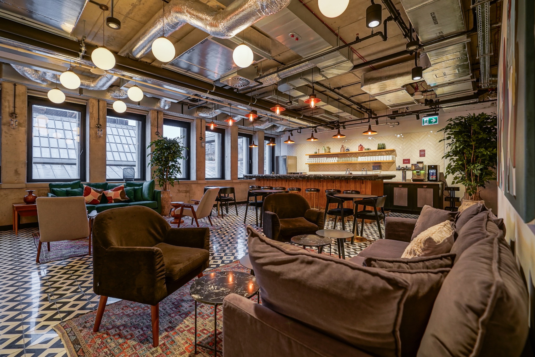 Przestronne wnętrze biurowe z sofami, fotelami i barem, oświetlone kulistymi lampami, z widocznymi elementami instalacji na suficie.