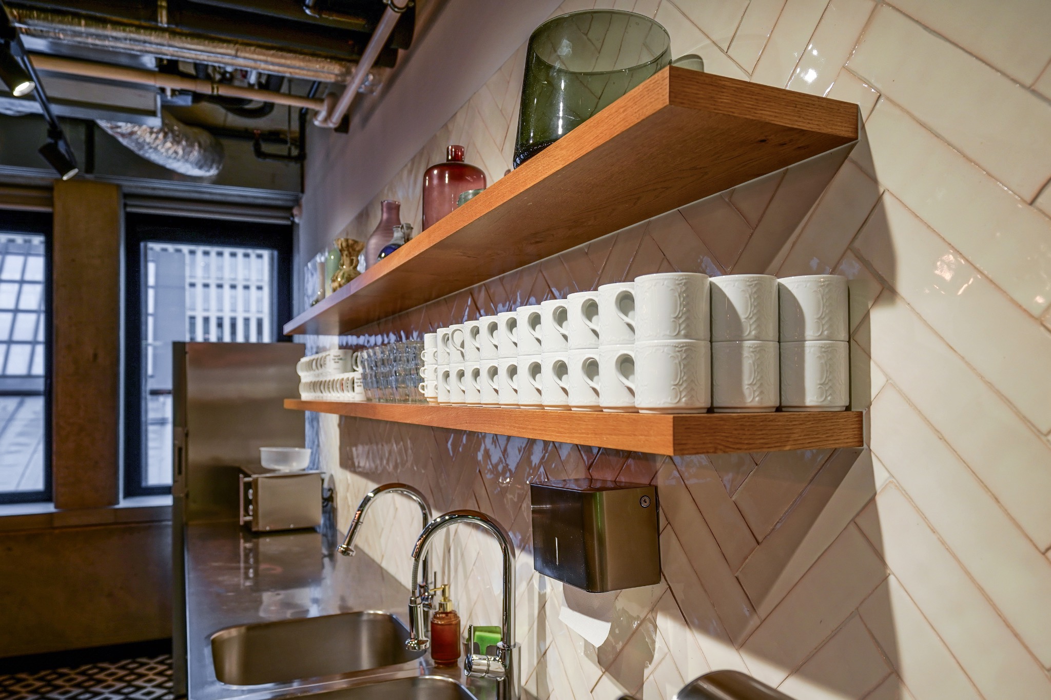 Biuro: Umywalka ze stali nierdzewnej, bateria, dozownik mydła i ręczników, na półkach drewnianych ułożone filiżanki i szklanki, tło stanowi ściana wyłożona błyszczącymi, kremowymi płytkami...