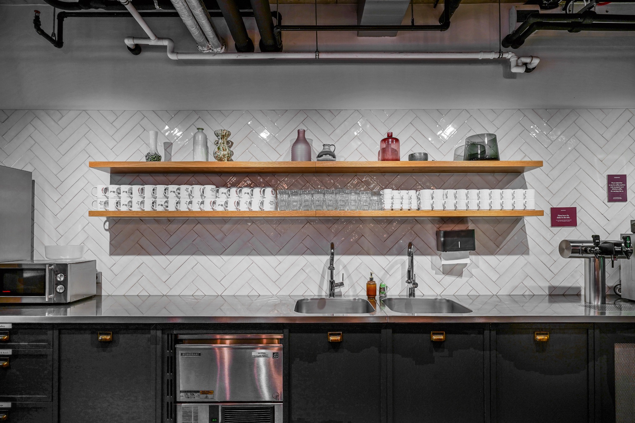 Nowoczesna kuchnia biurowa z dwoma zlewozmywakami, bateriami, półkami z naczyniami, lodówką i mikrofalówką na tle białych płytek ułożonych w jodełkę.