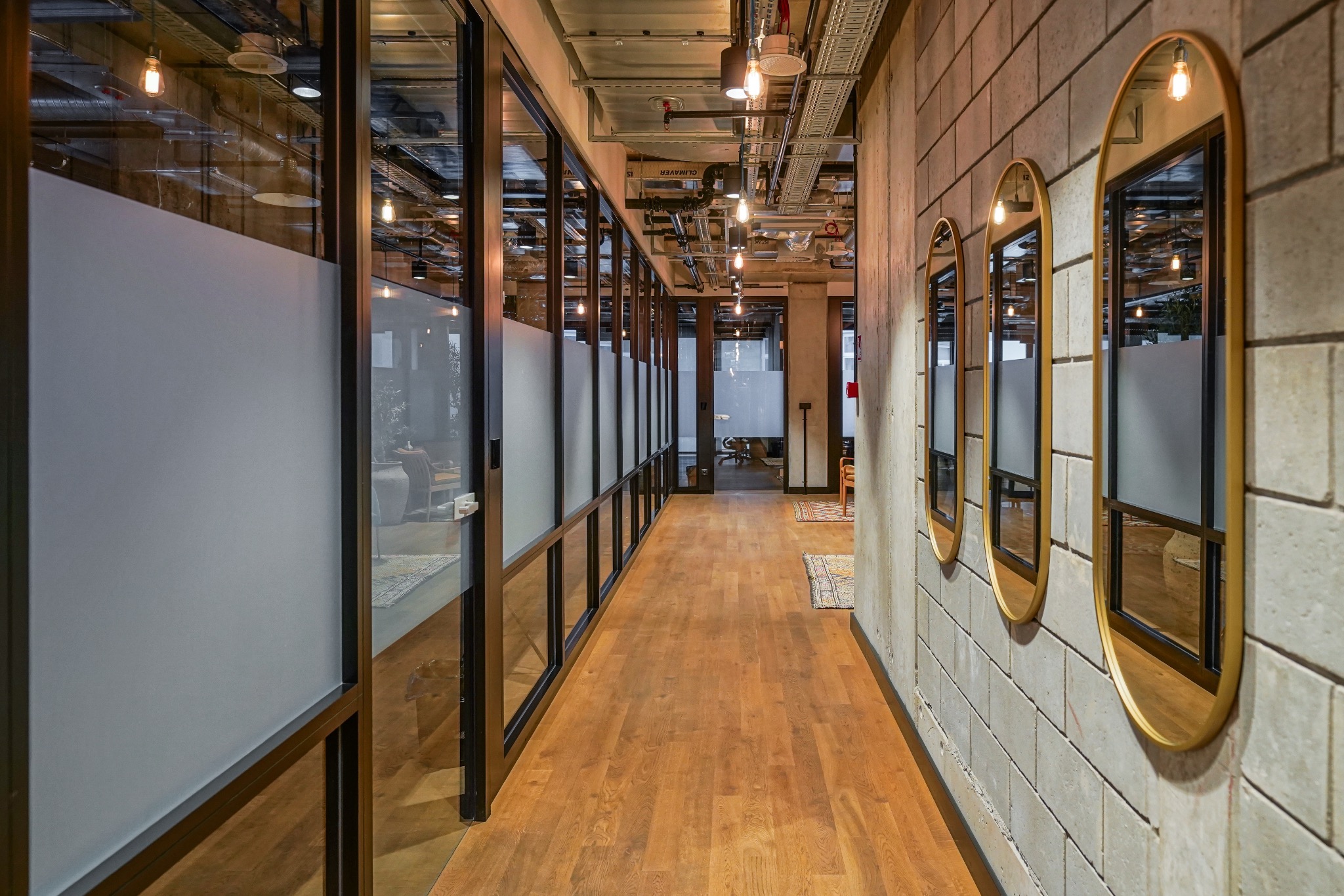 Korytarz biurowy z drewnianą podłogą, przeszklonymi ścianami z matowymi szybami, betonową ścianą z trzema owalnymi lustrami w złotych ramach i widocznymi instalacjami pod sufitem.