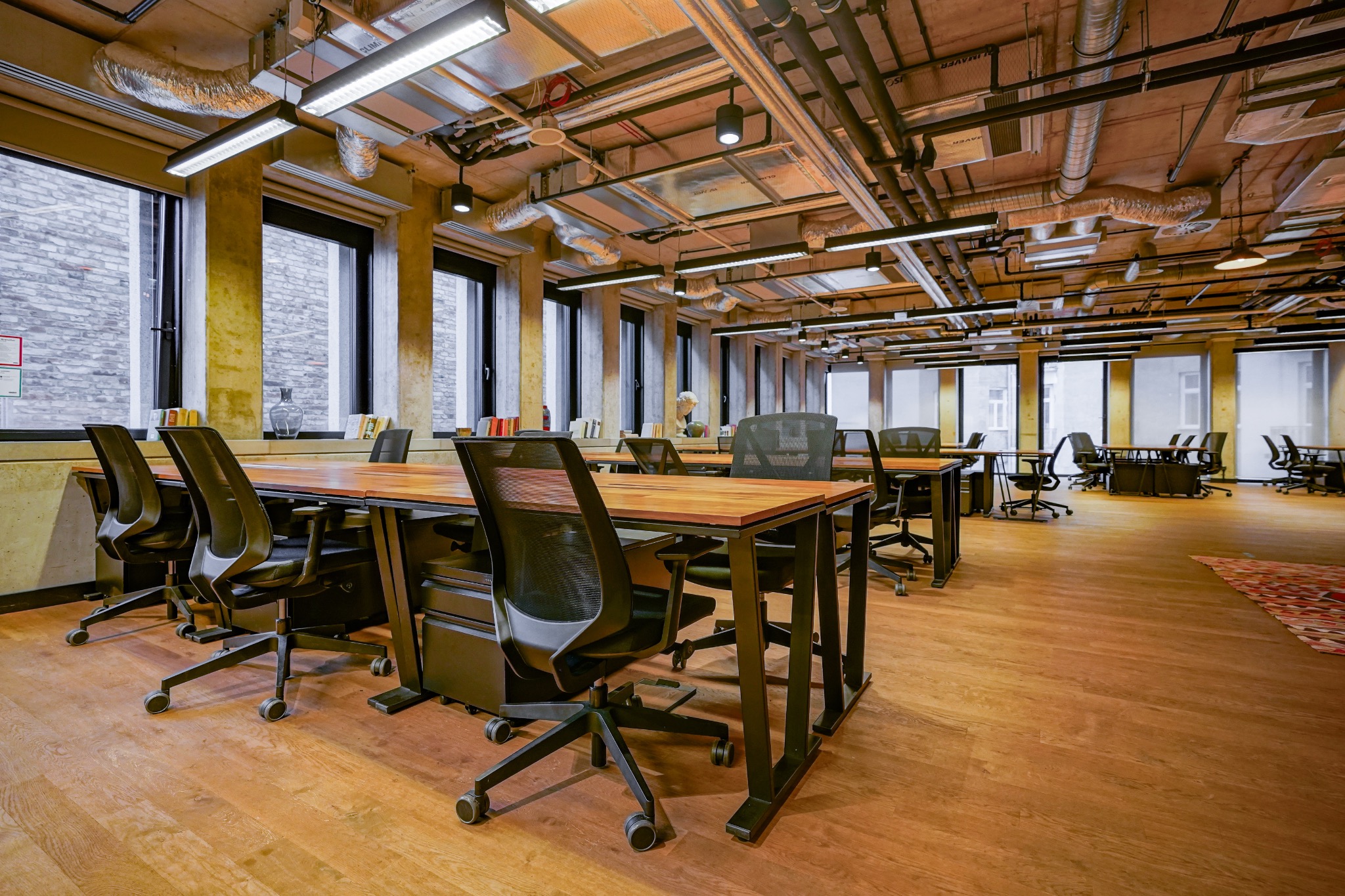 Przestronne biuro z drewnianą podłogą, rzędami biurek z czarnymi krzesłami, dużymi oknami i widocznymi instalacjami na suficie.
