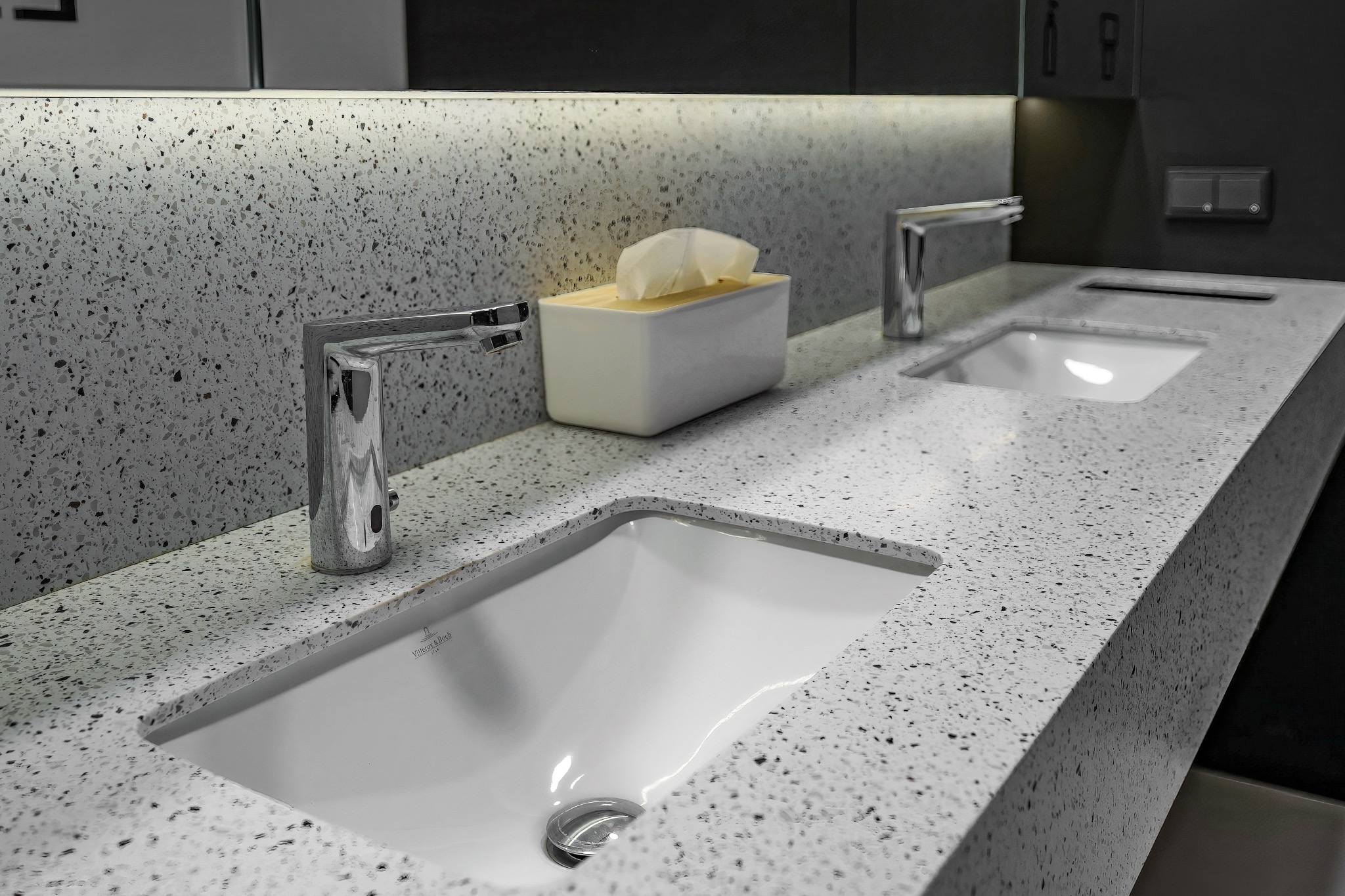 Biała umywalka Villeroy & Boch wkomponowana w blat z szarego lastryko, chromowana bateria, dozownik na chusteczki higieniczne w tle, oświetlenie LED pod szafką.