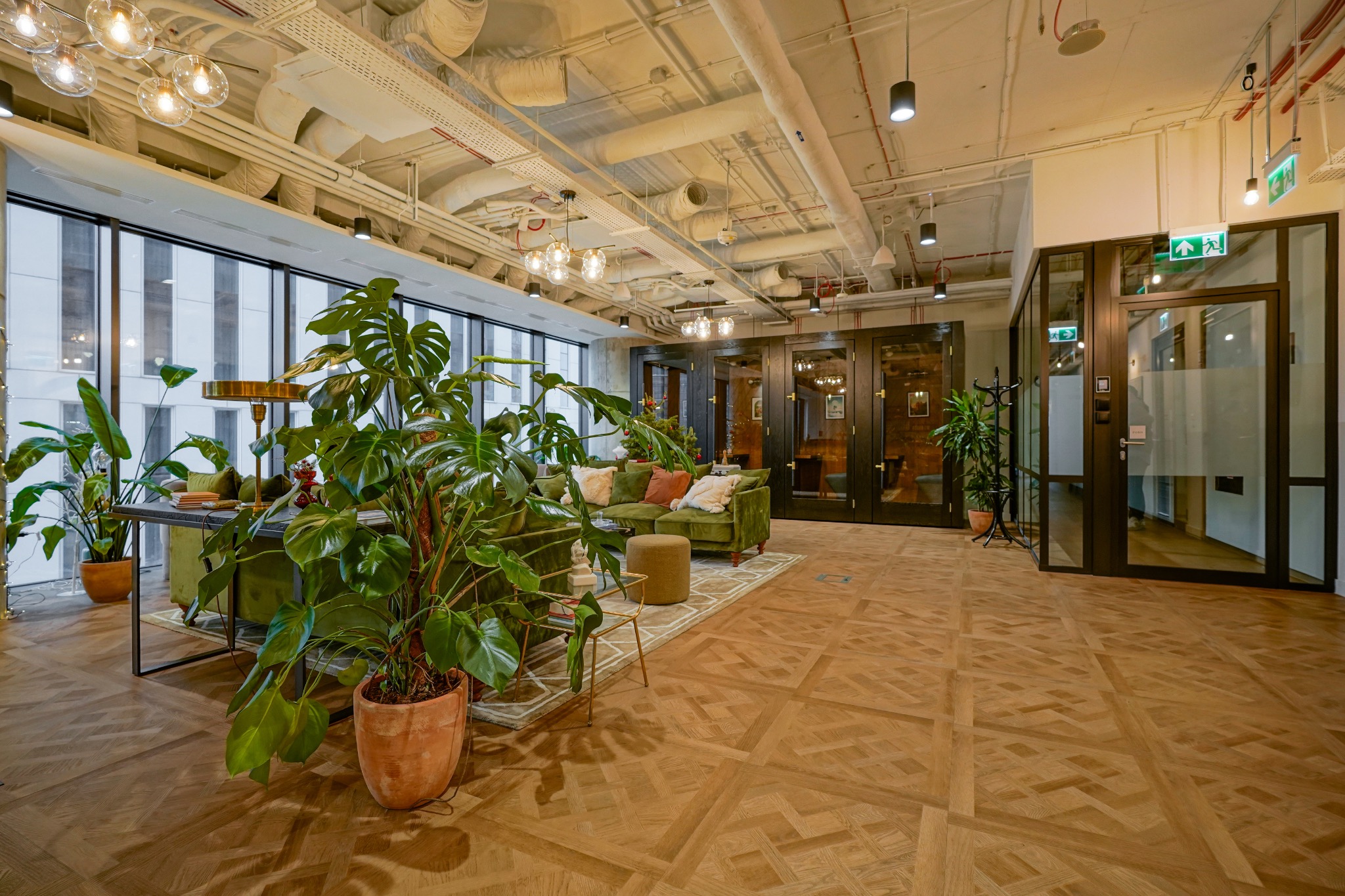 Jasne, przestronne biuro z parkietem w jodełkę, roślinami doniczkowymi, zieloną sofą i widocznymi instalacjami pod sufitem.