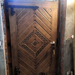 Piotr Chomiuk - Drewniane drzwi z górnym oknem łukowym, zdobione wzorem w romby i metalowymi okuciami, zamontowane w surowym murze z cegły i betonu.