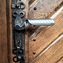 Piotr Chomiuk - Metalowa klamka z ozdobnym, kutym szyldem na drewnianych drzwiach z ukośnymi panelami i czarnymi nitami.