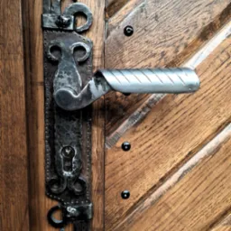 Metalowa klamka z ozdobnym, kutym szyldem na drewnianych drzwiach z ukośnymi panelami i czarnymi nitami.