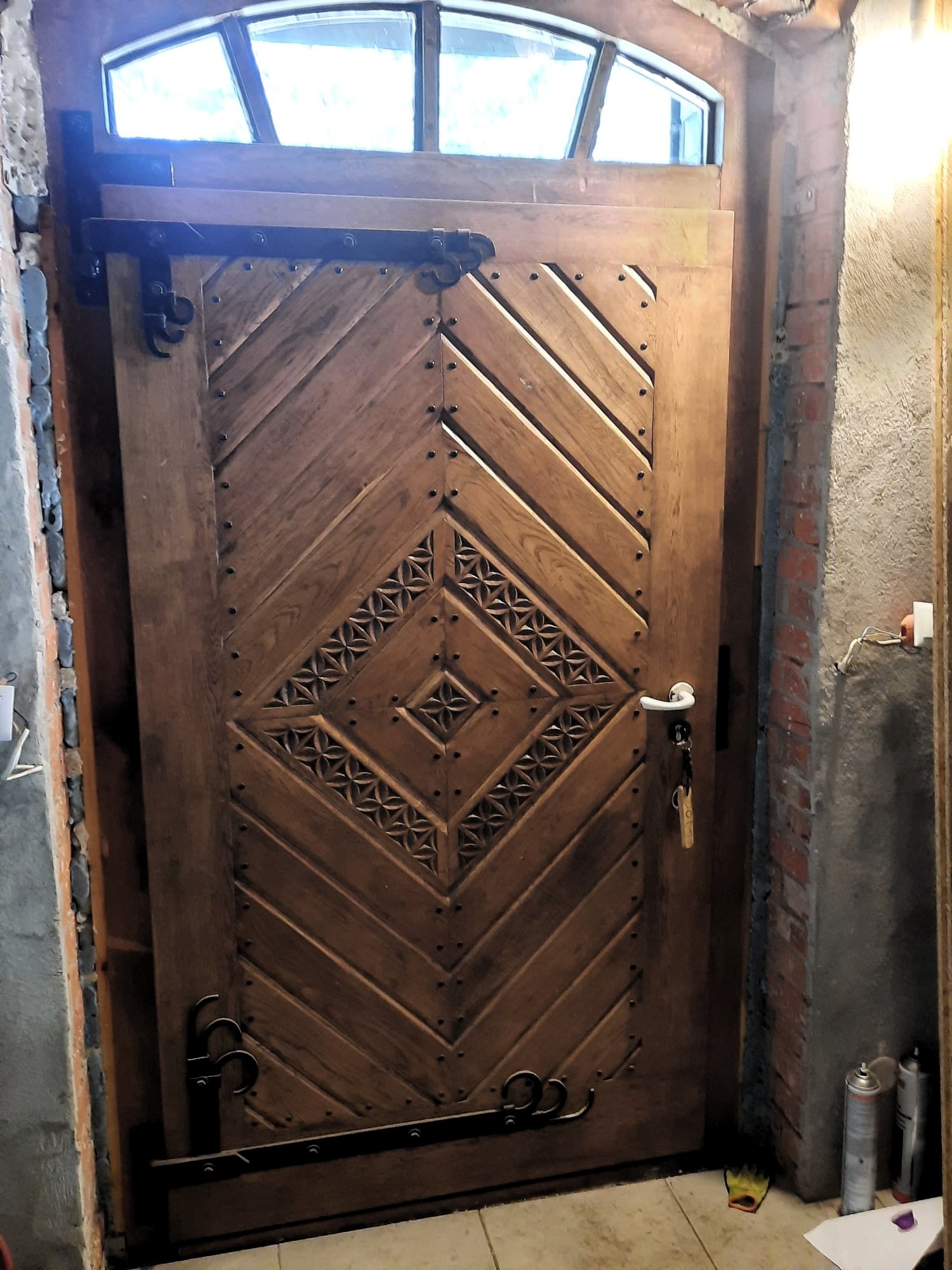 Drewniane drzwi z górnym oknem łukowym, zdobione wzorem w romby i metalowymi okuciami, zamontowane w surowym murze z cegły i betonu.