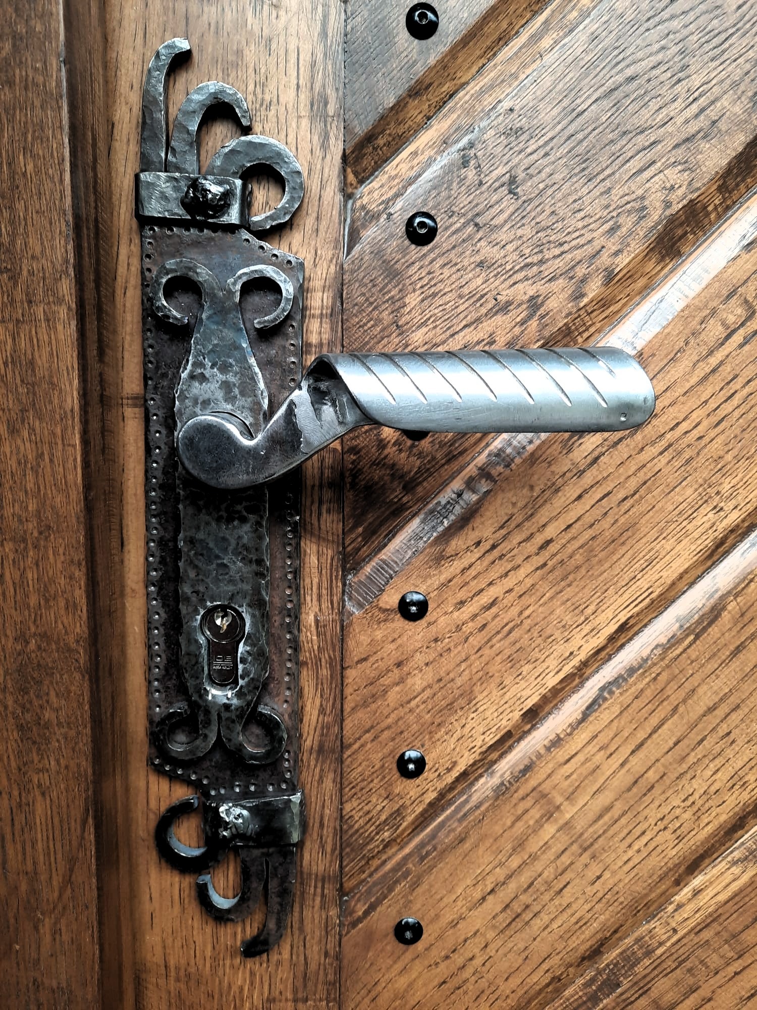 Metalowa klamka z ozdobnym, kutym szyldem na drewnianych drzwiach z ukośnymi panelami i czarnymi nitami.