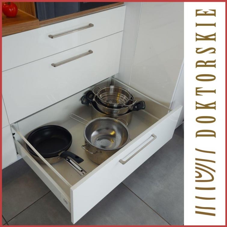 Biała szuflada kuchenna z pełnym wysuwem, zawierająca czarny rondel, metalowy durszlak i zestaw garnków.