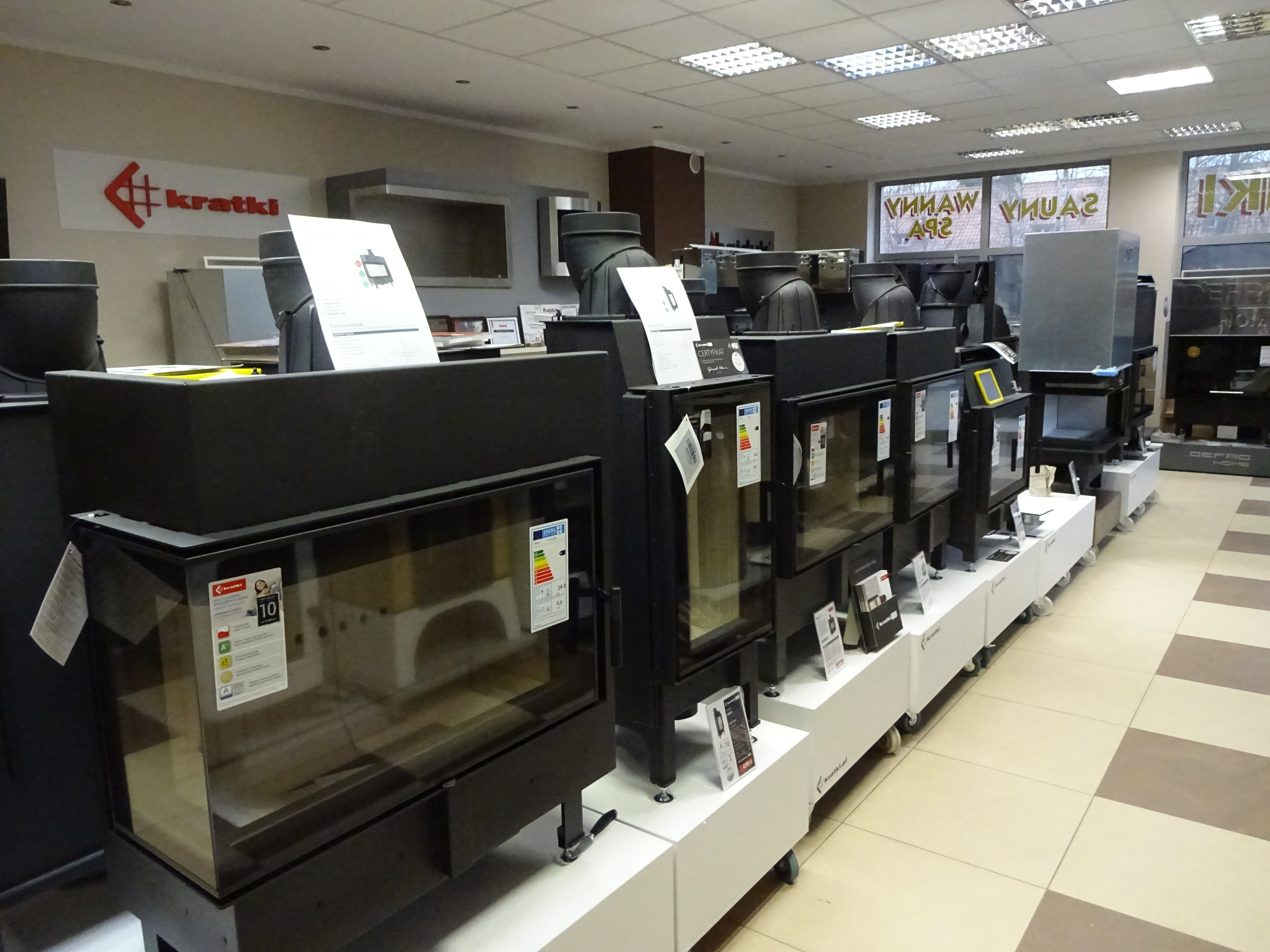 Ekspozycja w Olsztynie: Rząd czarnych, szklanych kominków marki Kratki na białych podestach w salonie. Widoczne etykiety energetyczne i logo producenta.