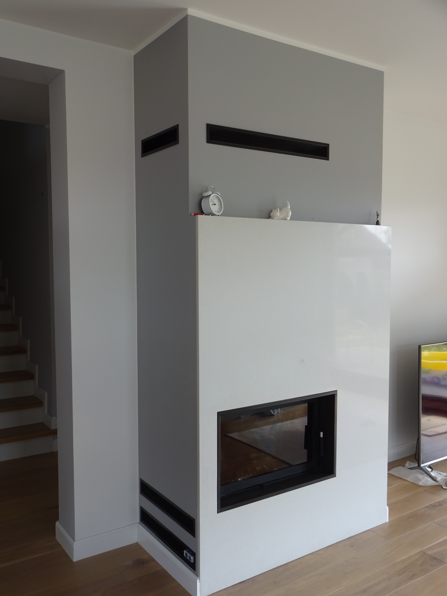 Nowoczesny kominek z białym obramowaniem i czarnymi wlotami powietrza, minimalistyczny design, zegar i figurka na górze, drewniana podłoga, szare ściany.