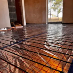 Mata grzewcza z czarnymi przewodami i czerwoną siatką ułożona na folii aluminiowej, widok z niskiego kąta w surowym wnętrzu z oknem na zewnątrz.