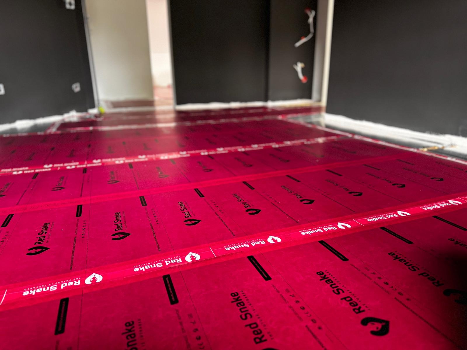 Czerwona folia grzewcza marki Red Snake rozłożona na podłodze w trakcie montażu, widoczne kable wystające ze ściany, przygotowanie do dalszych prac wykończeniowych.