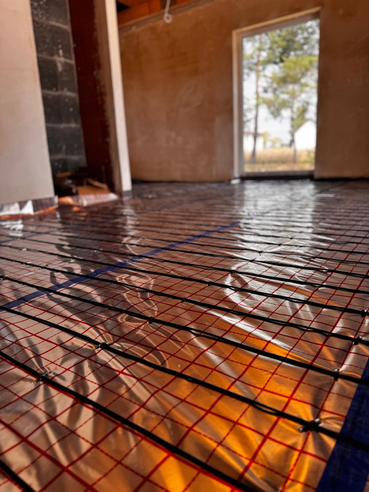 Mata grzewcza z czarnymi przewodami i czerwoną siatką ułożona na folii aluminiowej, widok z niskiego kąta w surowym wnętrzu z oknem na zewnątrz.