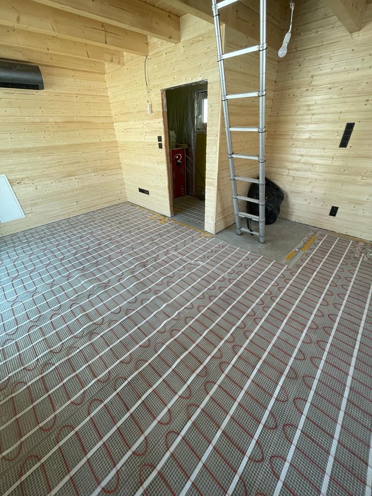 Wnętrze domu z bali w trakcie instalacji ogrzewania podłogowego: widoczne maty grzewcze z czerwonymi przewodami na podłożu, aluminiowa drabina oparta o ścianę, klimatyzator na ścianie z drewnianych...