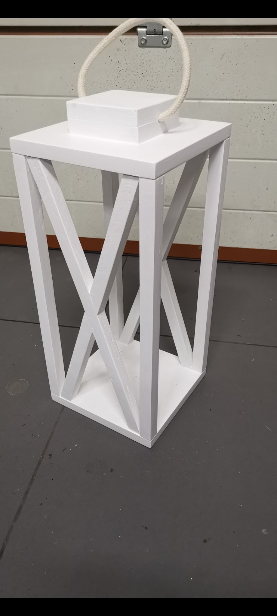 Biała, drewniana latarnia dekoracyjna z uchwytem z liny, ustawiona na szarym podłożu. Nowoczesny design z krzyżowymi elementami konstrukcyjnymi.