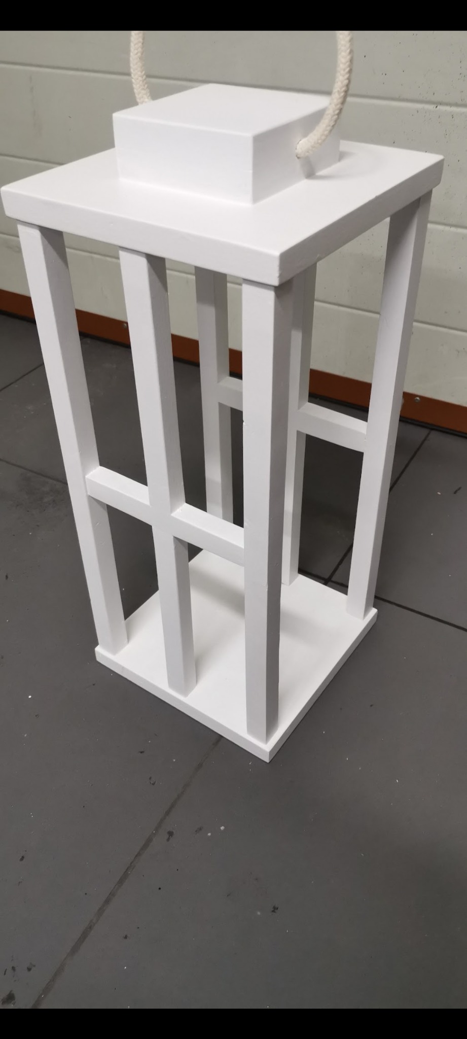 Biała, drewniana latarnia z uchwytem z liny, o geometrycznej konstrukcji, stojąca na szarym podłożu. Minimalistyczny design, widok z dołu.